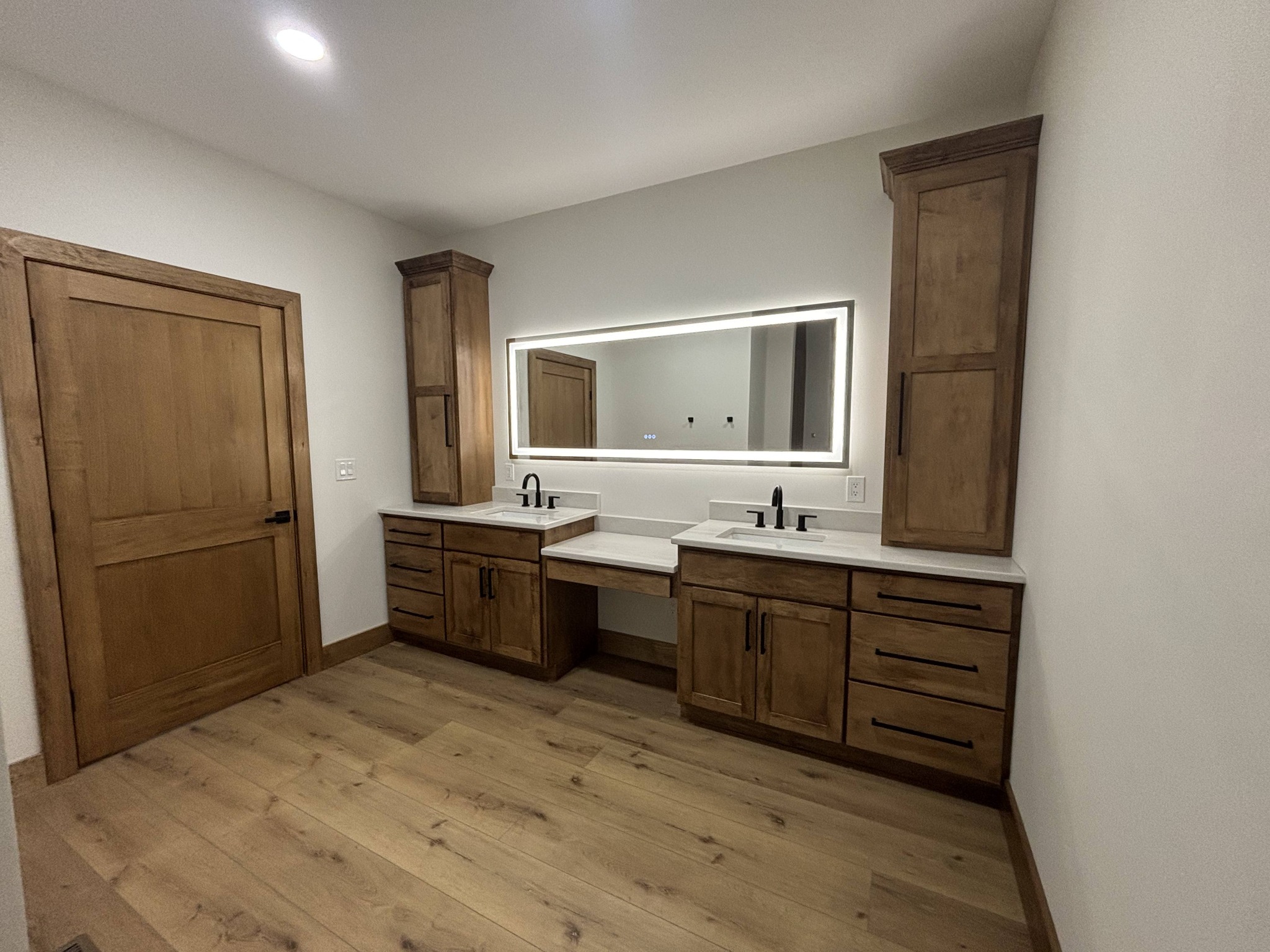 Spacious bathroom with wood cabinetry and dual sinks. A large mirror with backlighting adds a modern touch. Warm earth tones create a cozy feel.