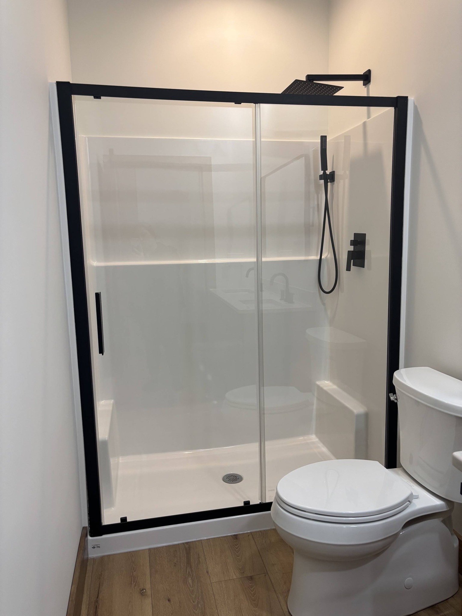 Modern bathroom with a sleek glass shower featuring black metal frames and a rain showerhead. White toilet, light wood floor, and minimalist design.