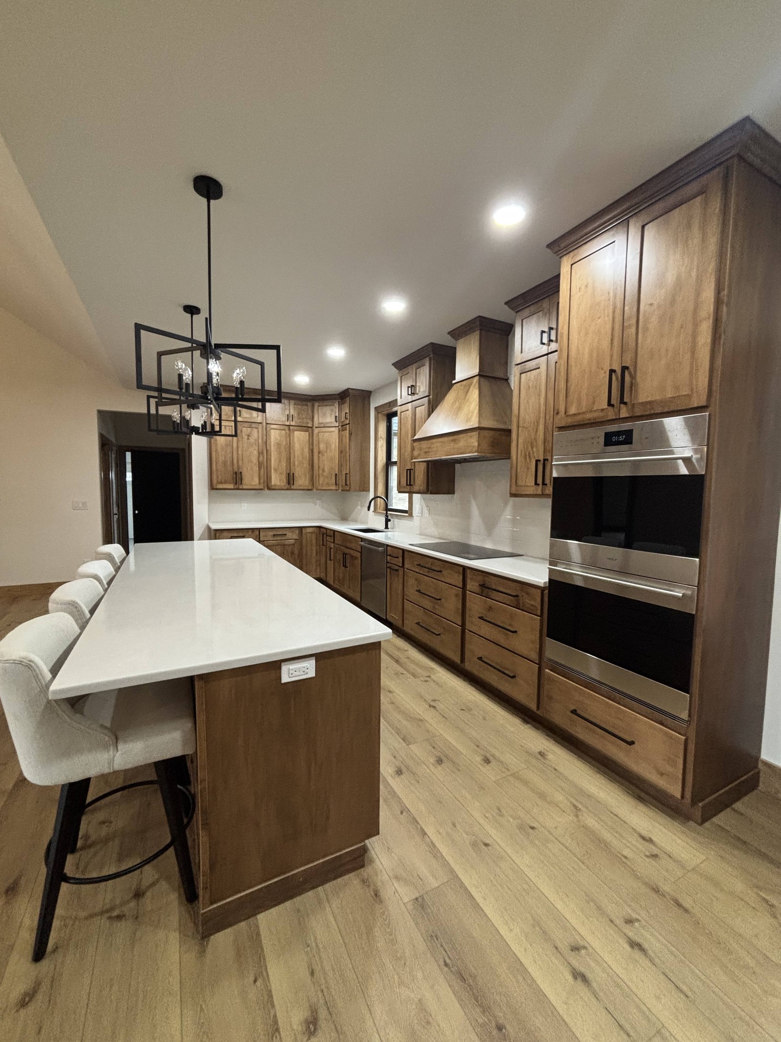 Modern kitchen with wooden cabinets and light wood floors. Features a large, white island with upholstered chairs and a black chandelier, creating a warm, inviting atmosphere.