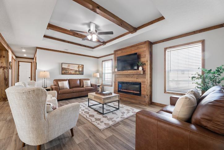 Spacious living room with brown leather sofas, beige armchairs, and a wood-accented wall with a TV and fireplace. Ceiling fan and large windows add light.