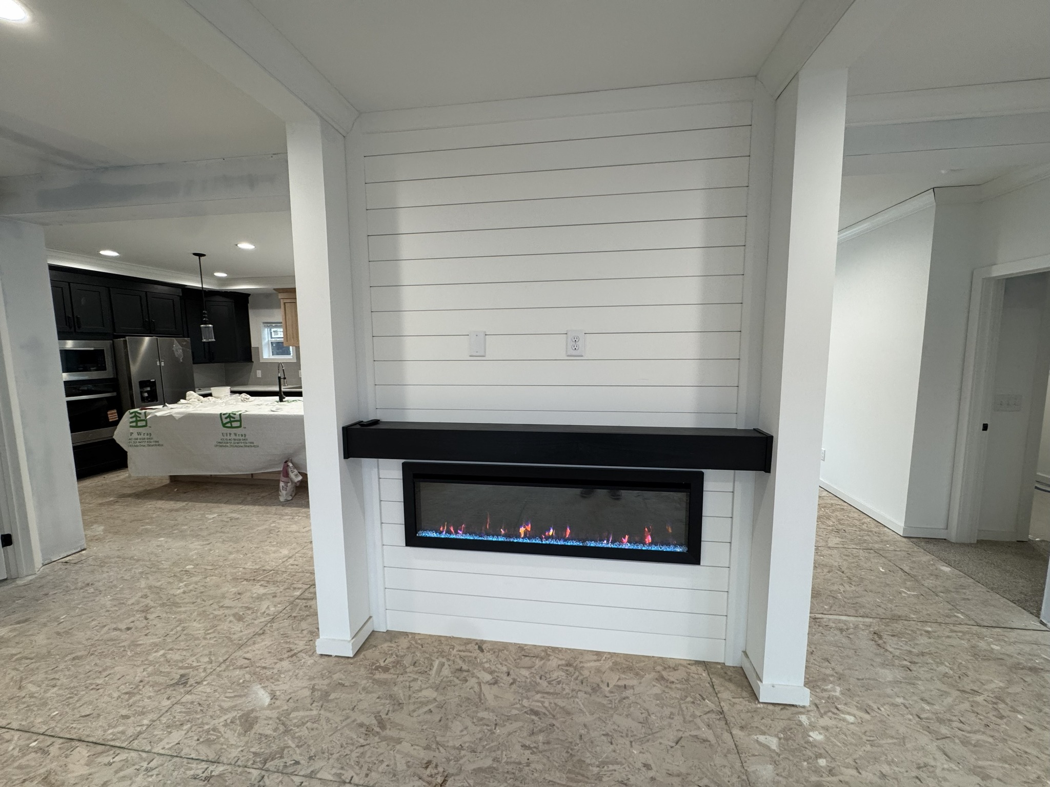 Modern interior with a minimalist fireplace set in a white shiplap wall. Visible are unfinished floors and a kitchen area with covered counters.