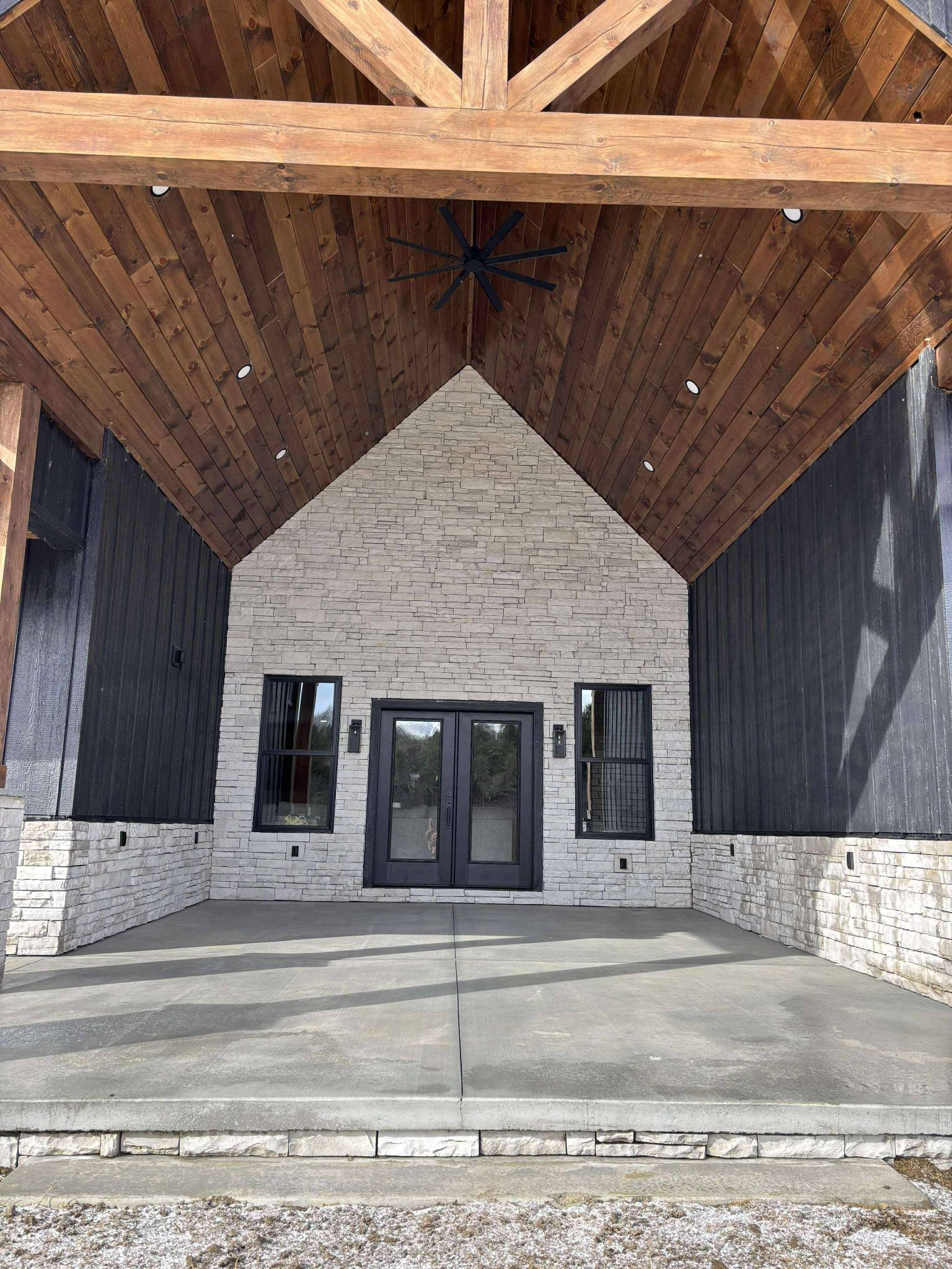 Rustic porch with a peaked wooden ceiling and stone walls, featuring double black-framed glass doors and two windows, creating a cozy, inviting atmosphere.