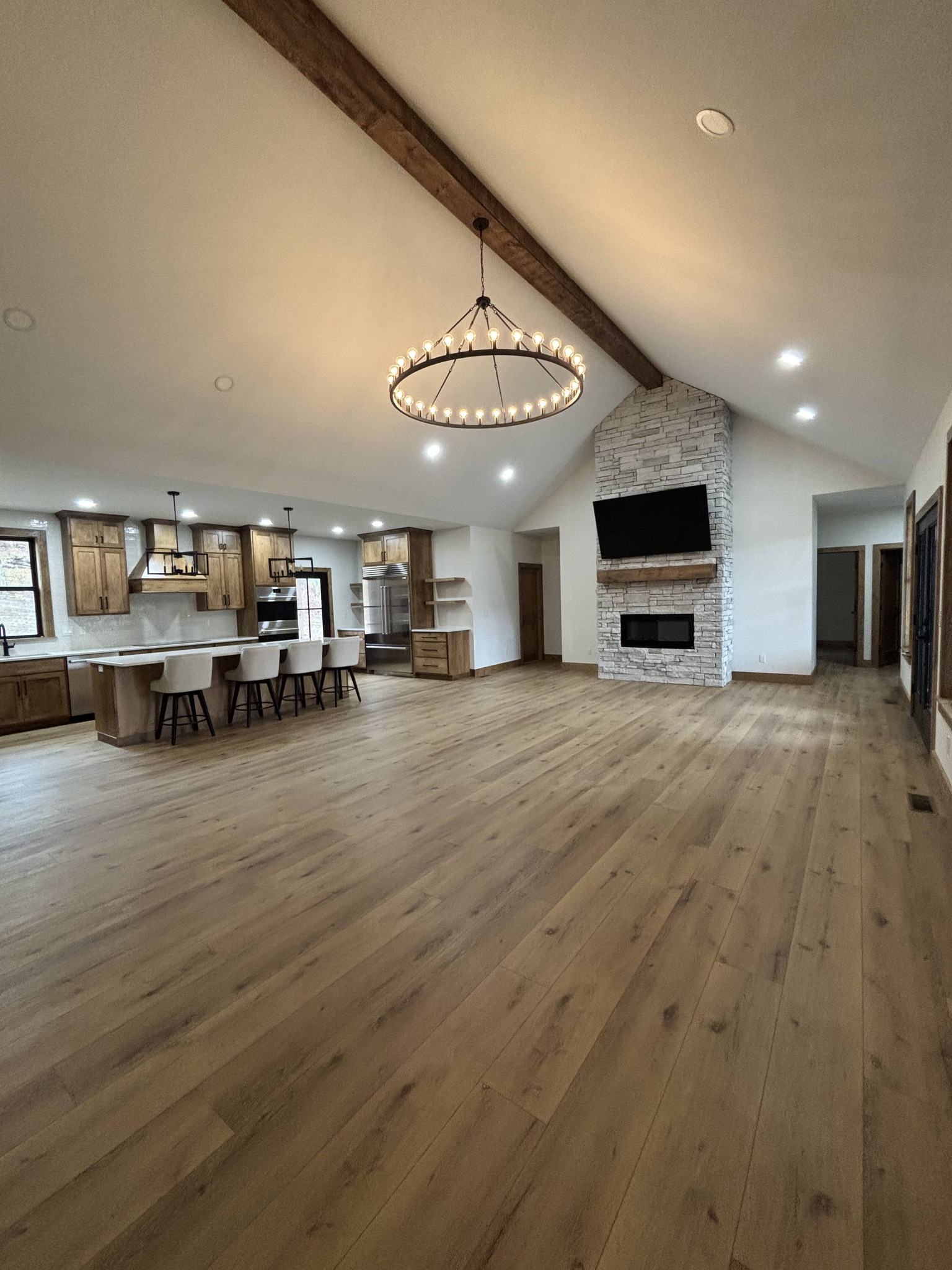 Spacious open-concept living area with wooden floors, large chandelier, modern kitchen with island, stone fireplace, and high vaulted ceiling.