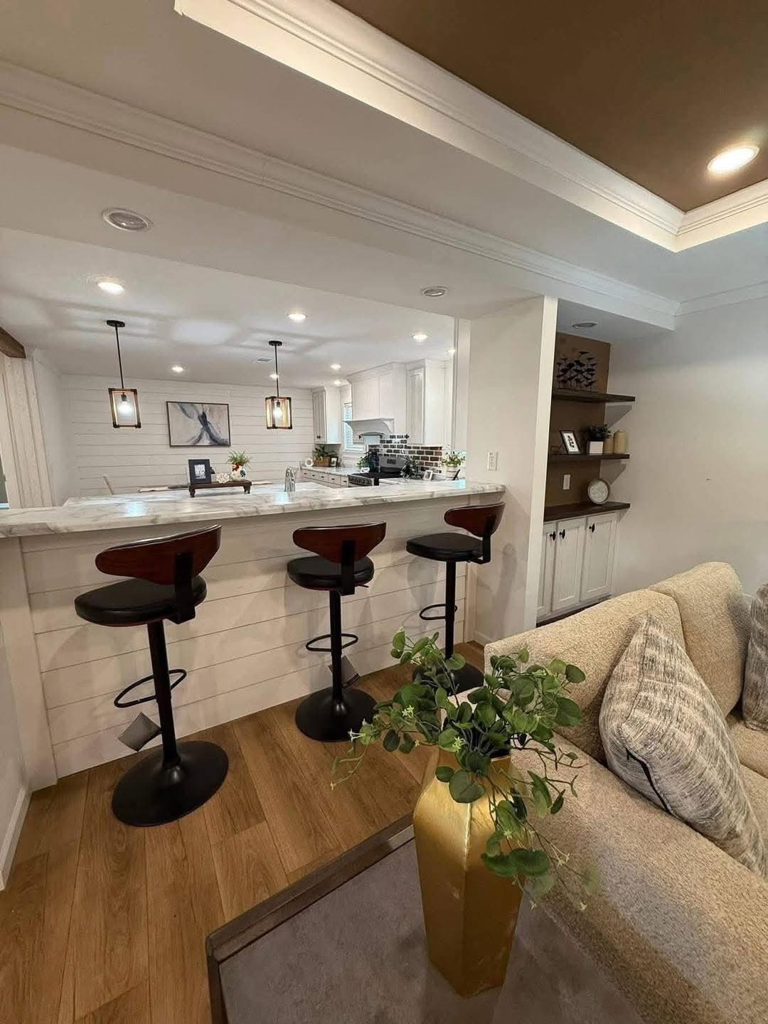 A cozy kitchen and living area featuring a wooden floor, marble countertop with three bar stools, pendant lighting, and a beige sofa with cushions. A warm, inviting atmosphere.