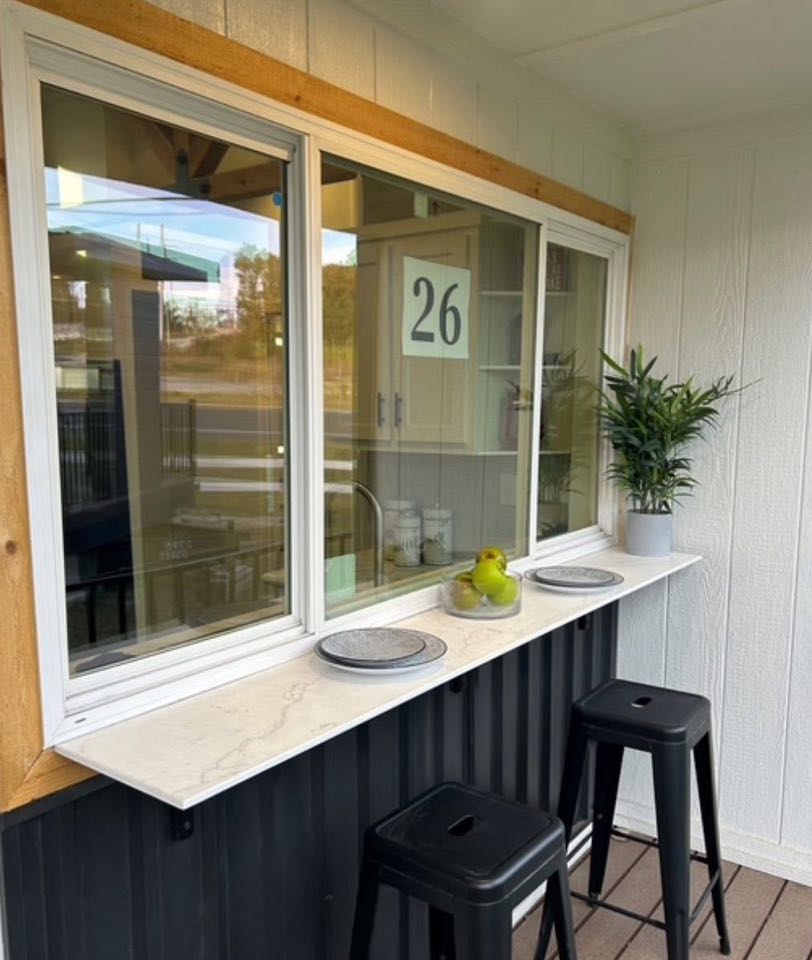 Outdoor bar-style countertop with two black stools in front of a window. Decor includes plates, a plant, and a fruit bowl. Bright and inviting setup.