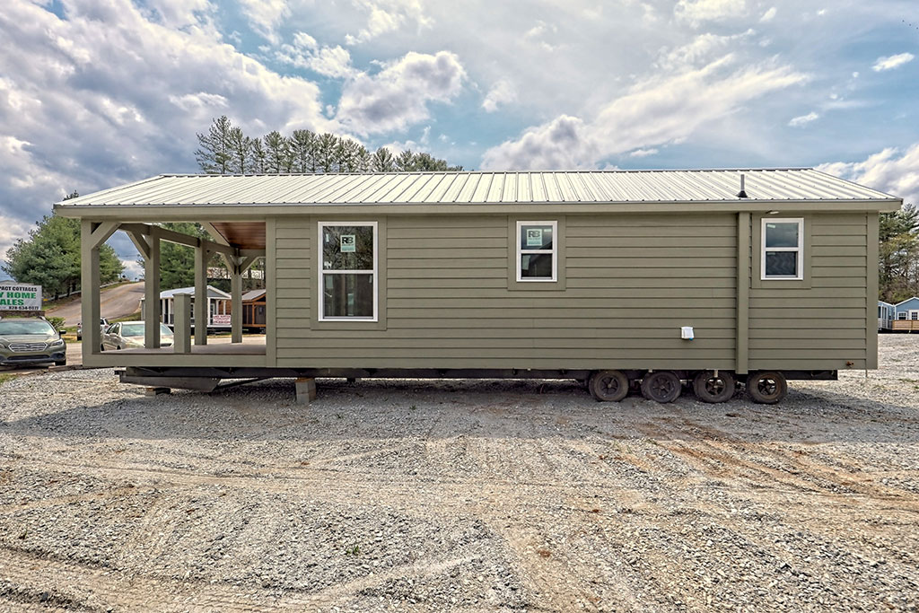 Single-story, beige tiny house on wheels sits on a gravel lot, under a partly cloudy sky. Minimalistic design with a cozy porch suggests a mobile home.
