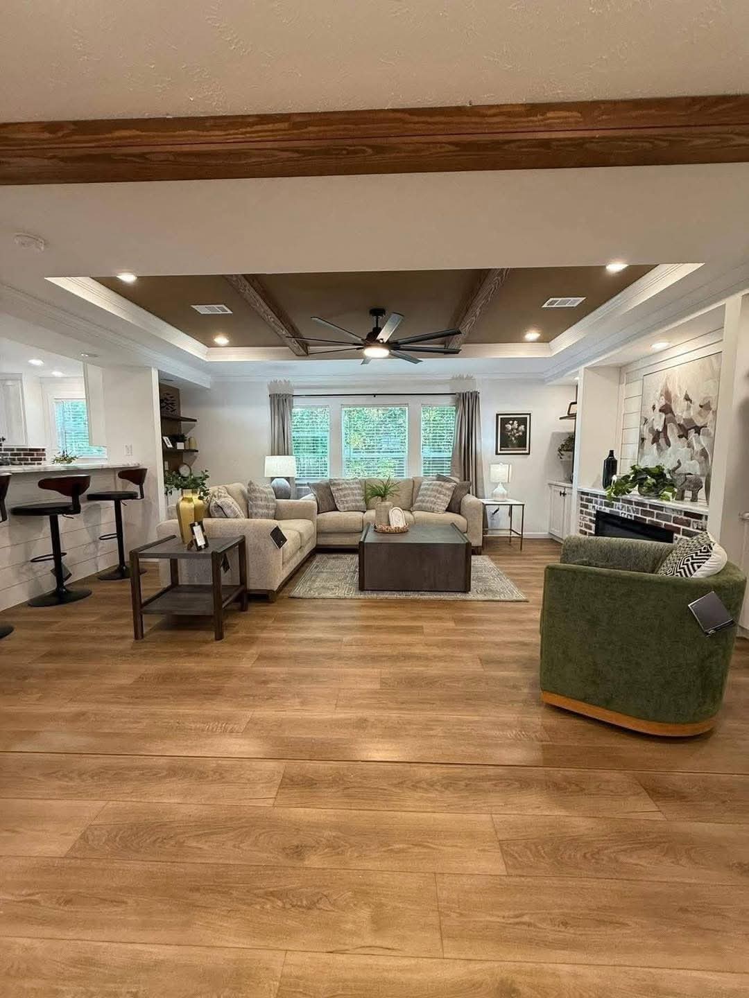 Cozy living room with wooden floors, beige couches, a central coffee table, and a ceiling fan. Natural light filters through large windows, creating a warm ambiance.