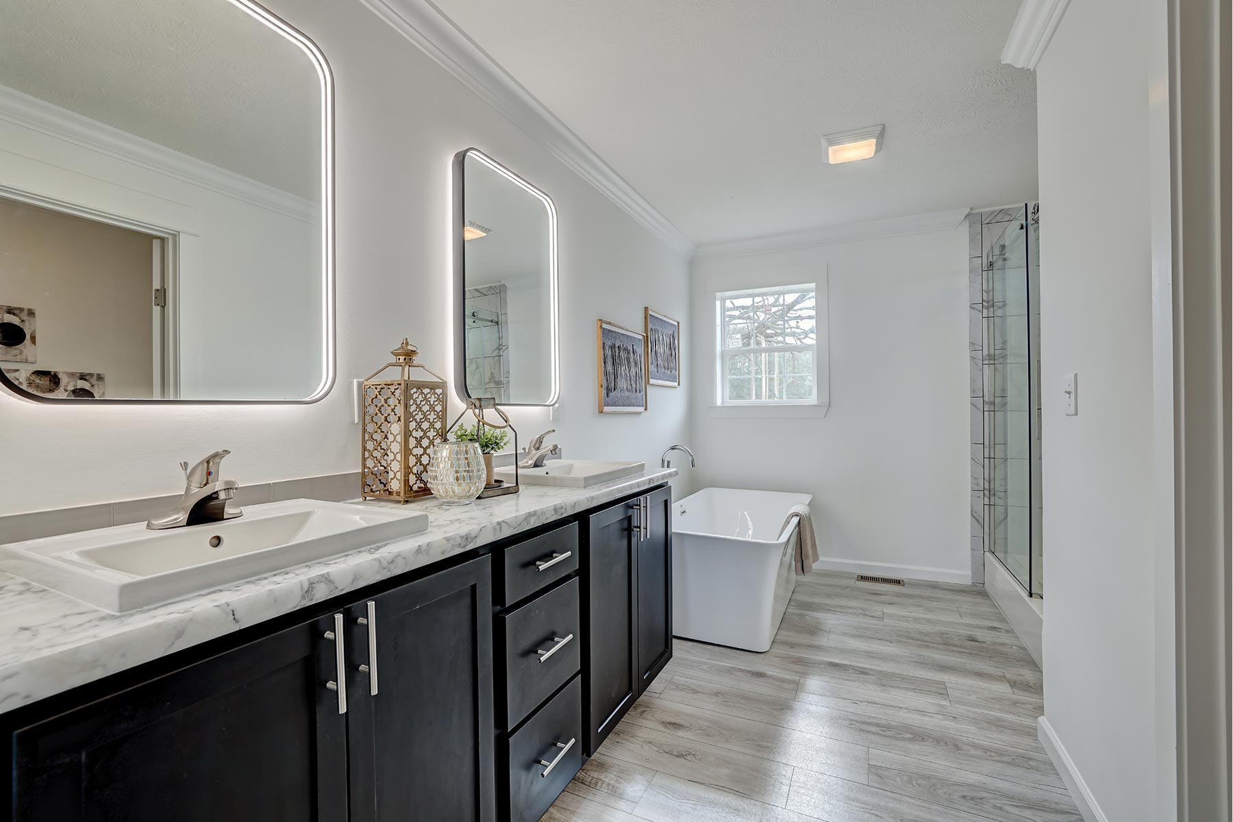 Modern bathroom with dark wood cabinets, illuminated mirrors, dual sinks, and marble countertops. A freestanding bathtub and glass shower sit beside a window.