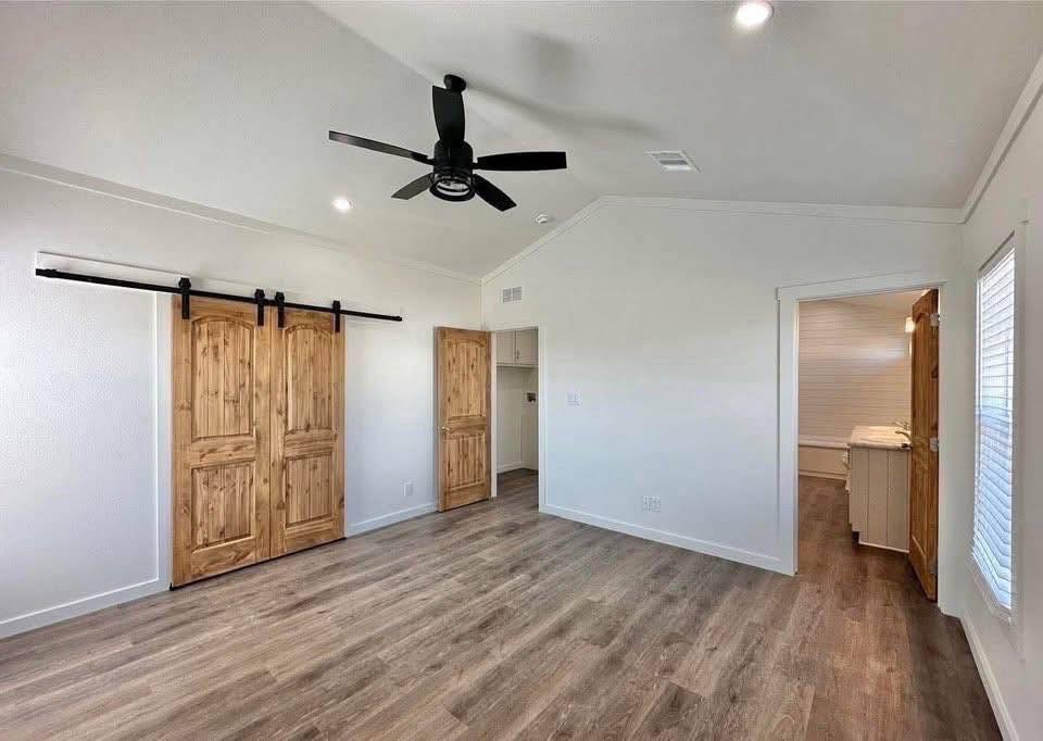 Spacious room with natural tones, featuring wooden barn doors, a dark ceiling fan, and sleek wooden flooring. An open doorway reveals a bright adjacent area.