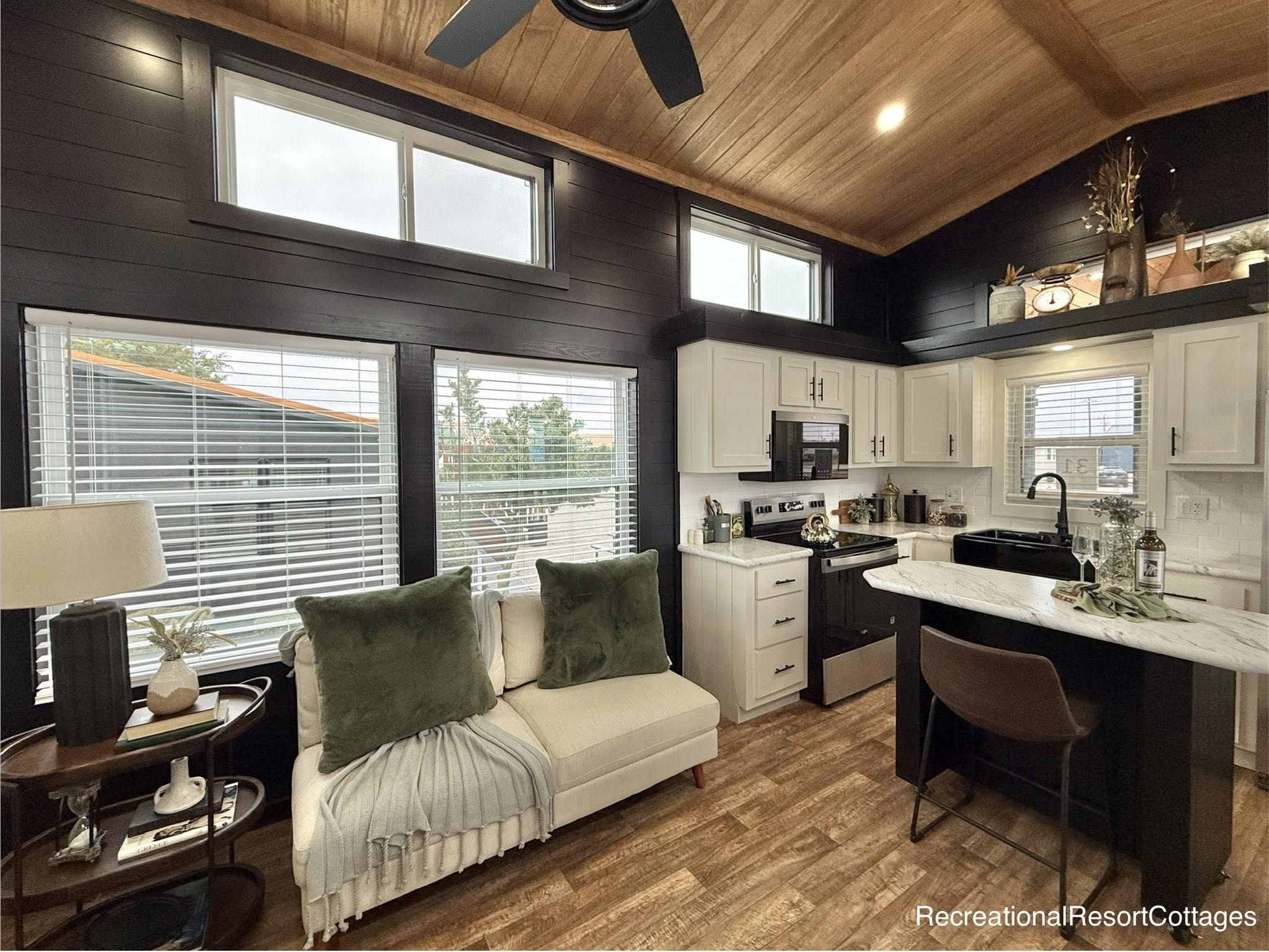 Cozy kitchen and living area in a small home features black and white decor, wooden ceiling, large windows, and plush seating, evoking warmth.