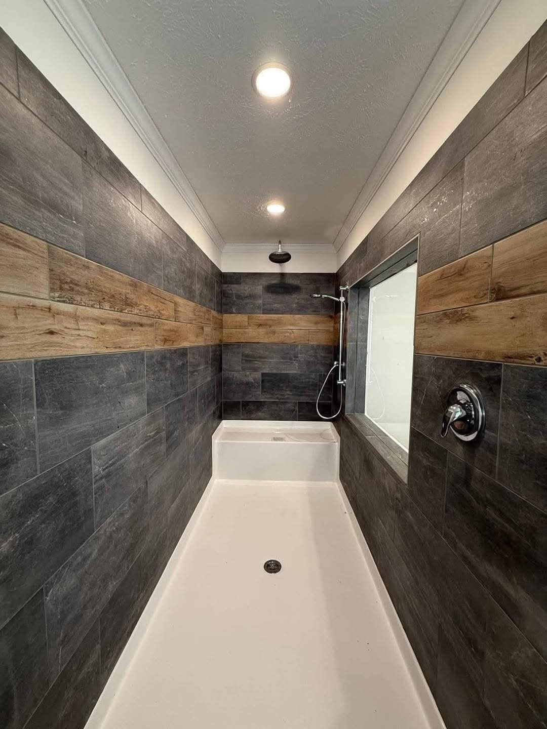 Modern shower with dark gray tiles and a wood accent strip, featuring a white base and ceiling lights enhancing a sleek, minimalist design.