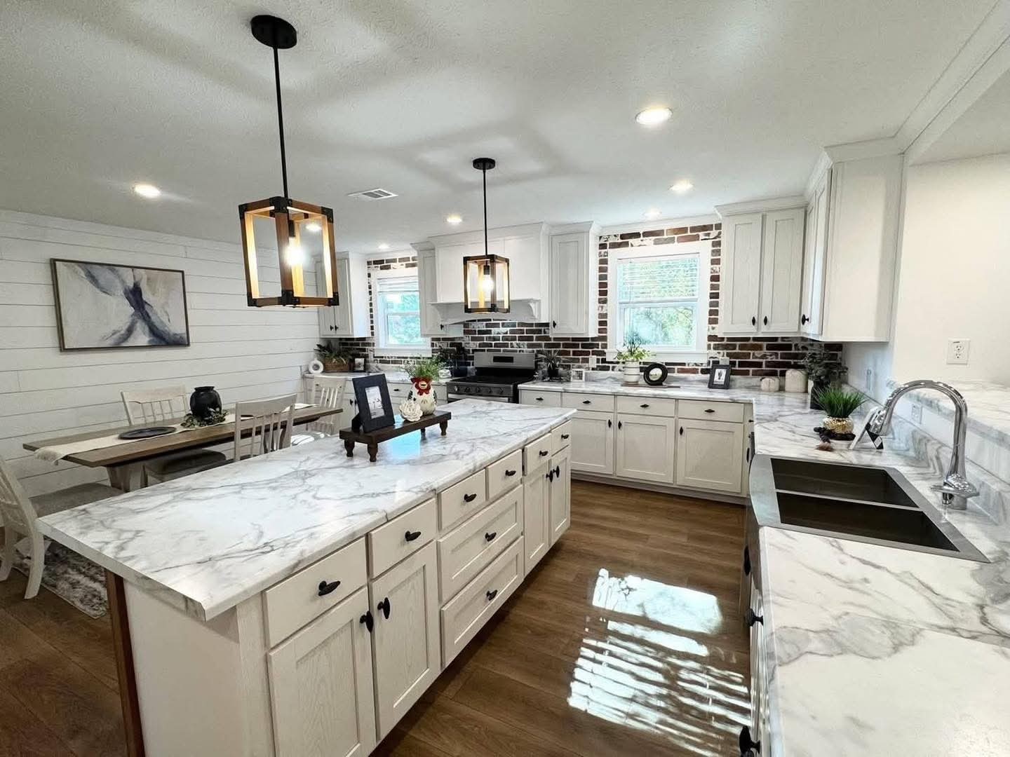 Bright kitchen with white cabinets and marble countertops. Two pendant lights hang above a large island. Brick backsplash adds rustic charm. Cozy dining area with wooden table on the side.