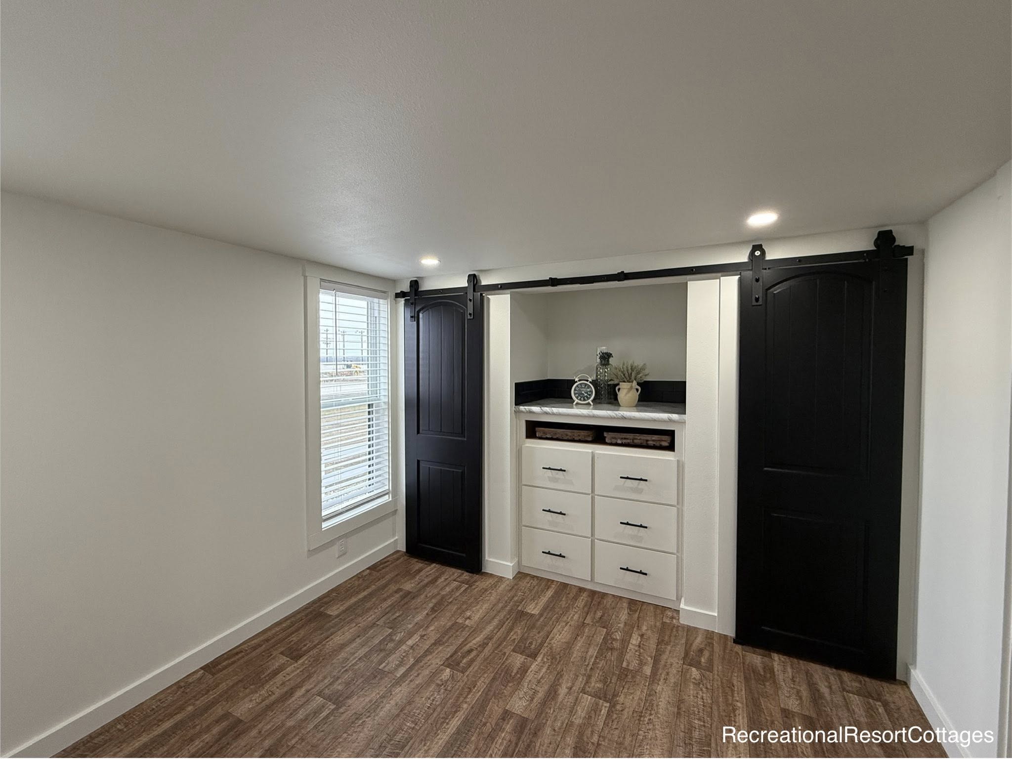 Cozy room with wood floor, white walls, and recessed lighting. A window with blinds is on the left. Centered are white drawers framed by two black sliding barn doors.