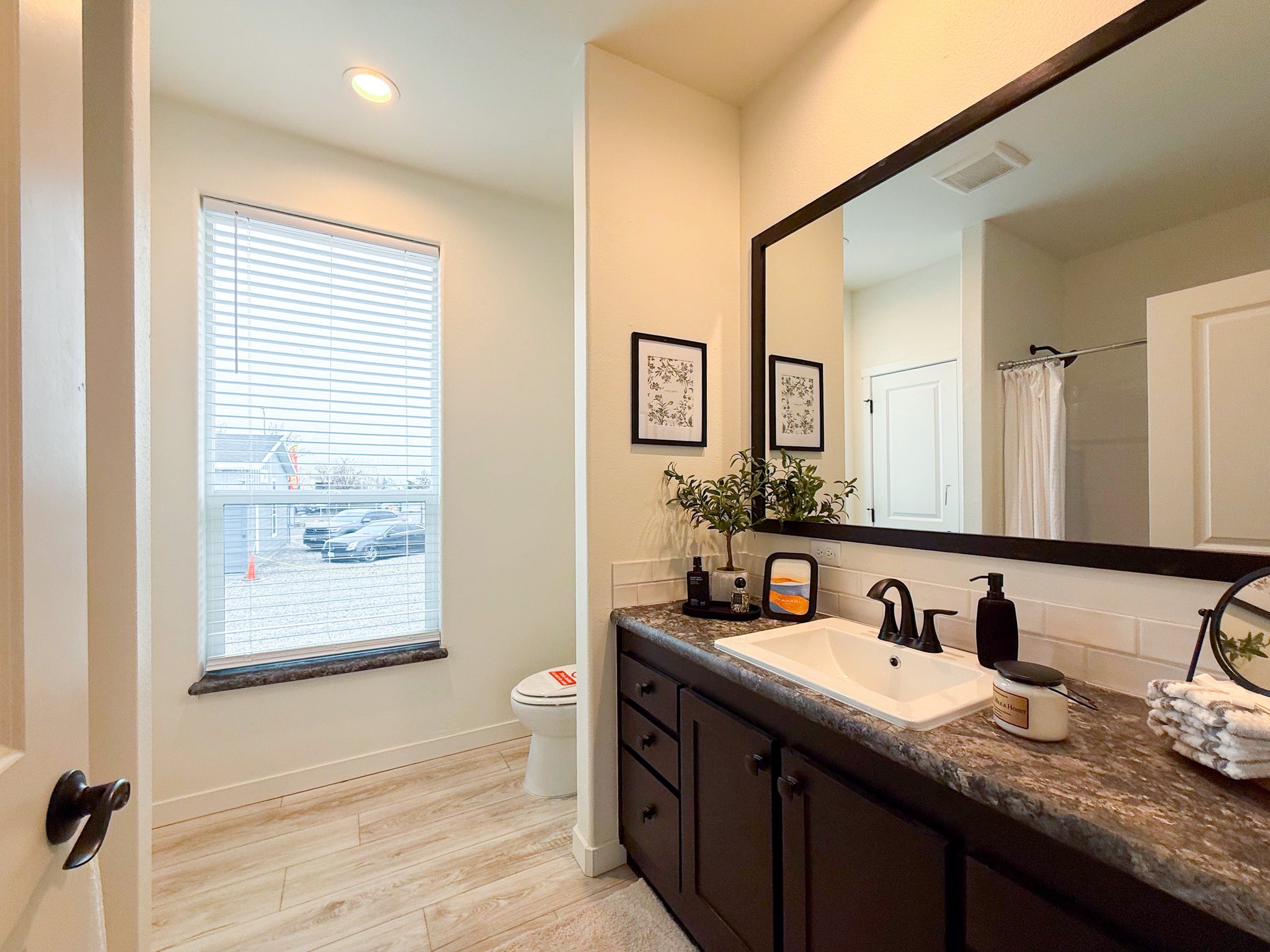 A bright, modern bathroom with a large mirror above a dark countertop, a white sink, plants, and decor. Light wood flooring and a window with blinds.