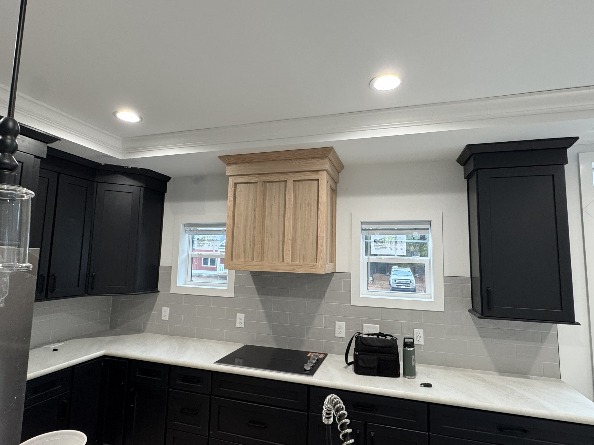 Modern kitchen with black cabinets, a light wooden range hood, and a sleek electric stovetop. Two small windows add natural light, creating a fresh ambiance.