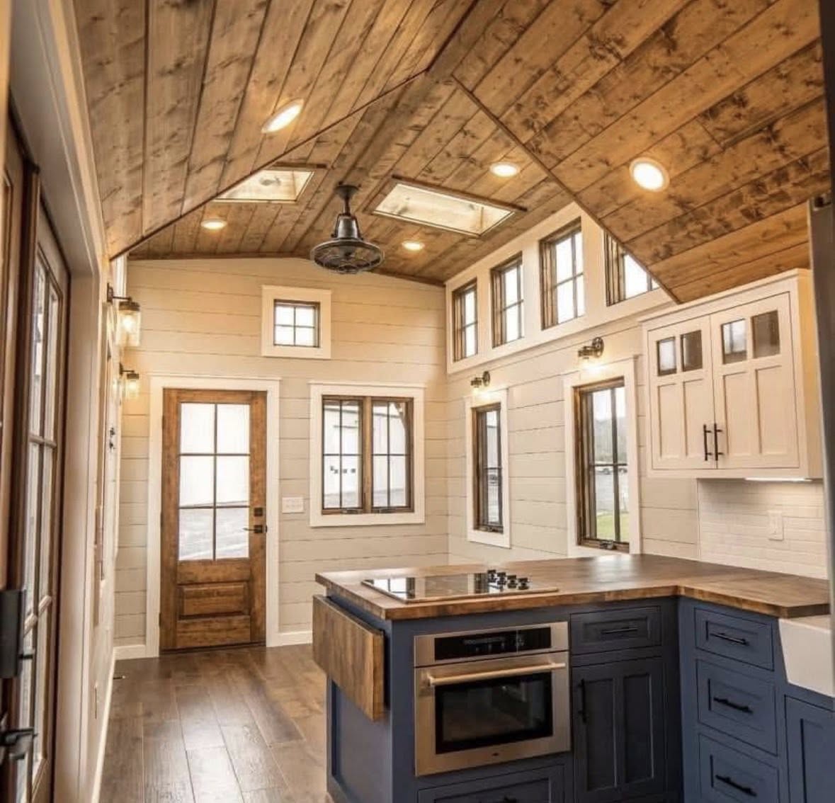 Cozy kitchen with vaulted wood ceiling, recessed lights, and skylights. White shiplap walls, rustic wood door, navy cabinets, and wooden countertops.