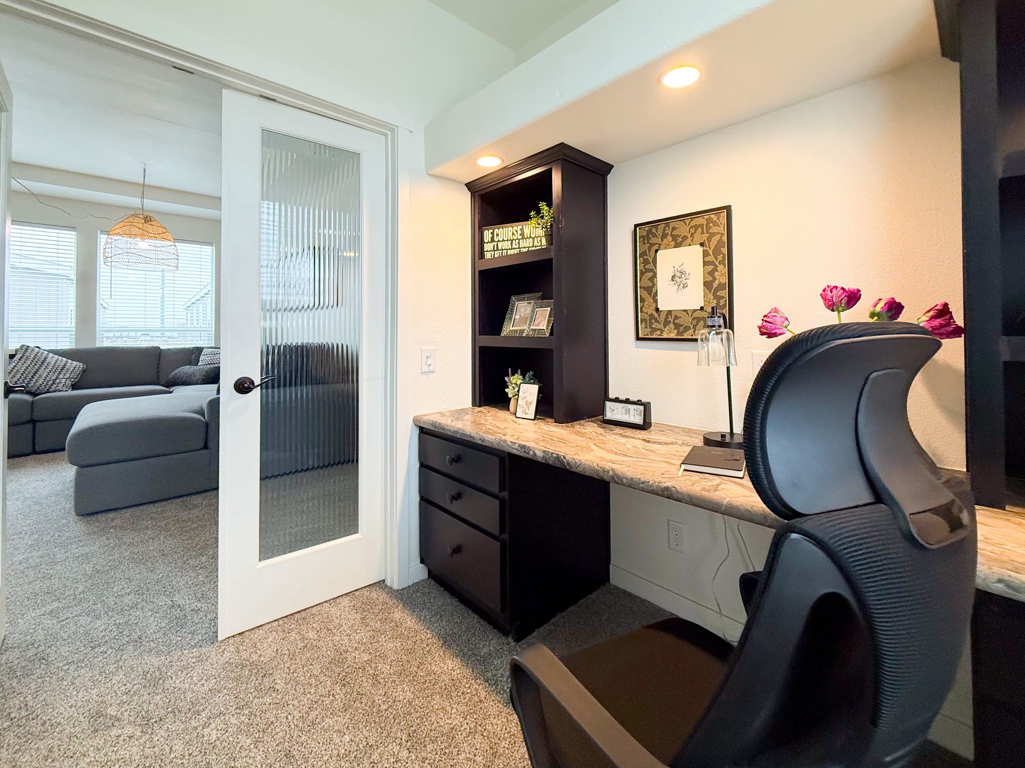 Modern home office with a marble desk, black chair, and dark shelving adorned with decor and framed art. A cozy living room with a gray couch is visible through a glass door.