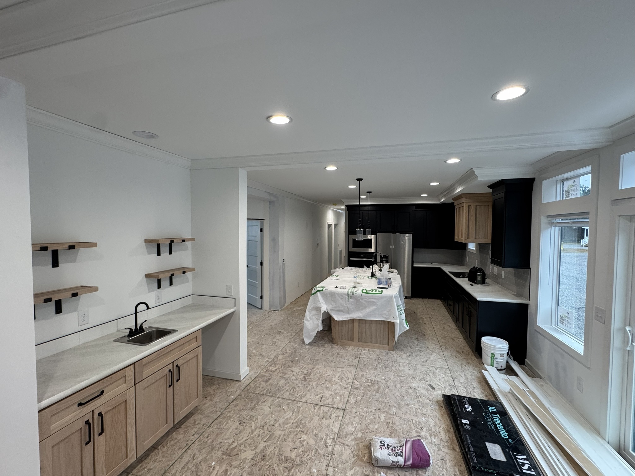 Modern kitchen under renovation with light wood cabinets, black appliances, and open shelving. Floor unfinished. Bright lighting creates a spacious feel.