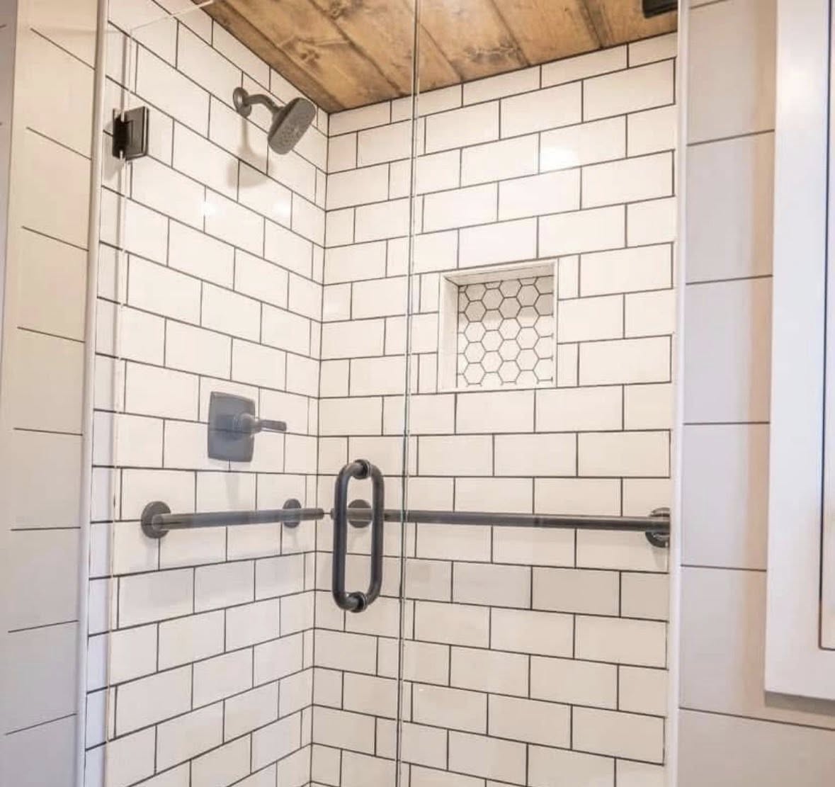 Modern shower with white subway tiles, glass door, and black fixtures. Wooden ceiling, small hexagonal tile niche. Clean and minimalist design.