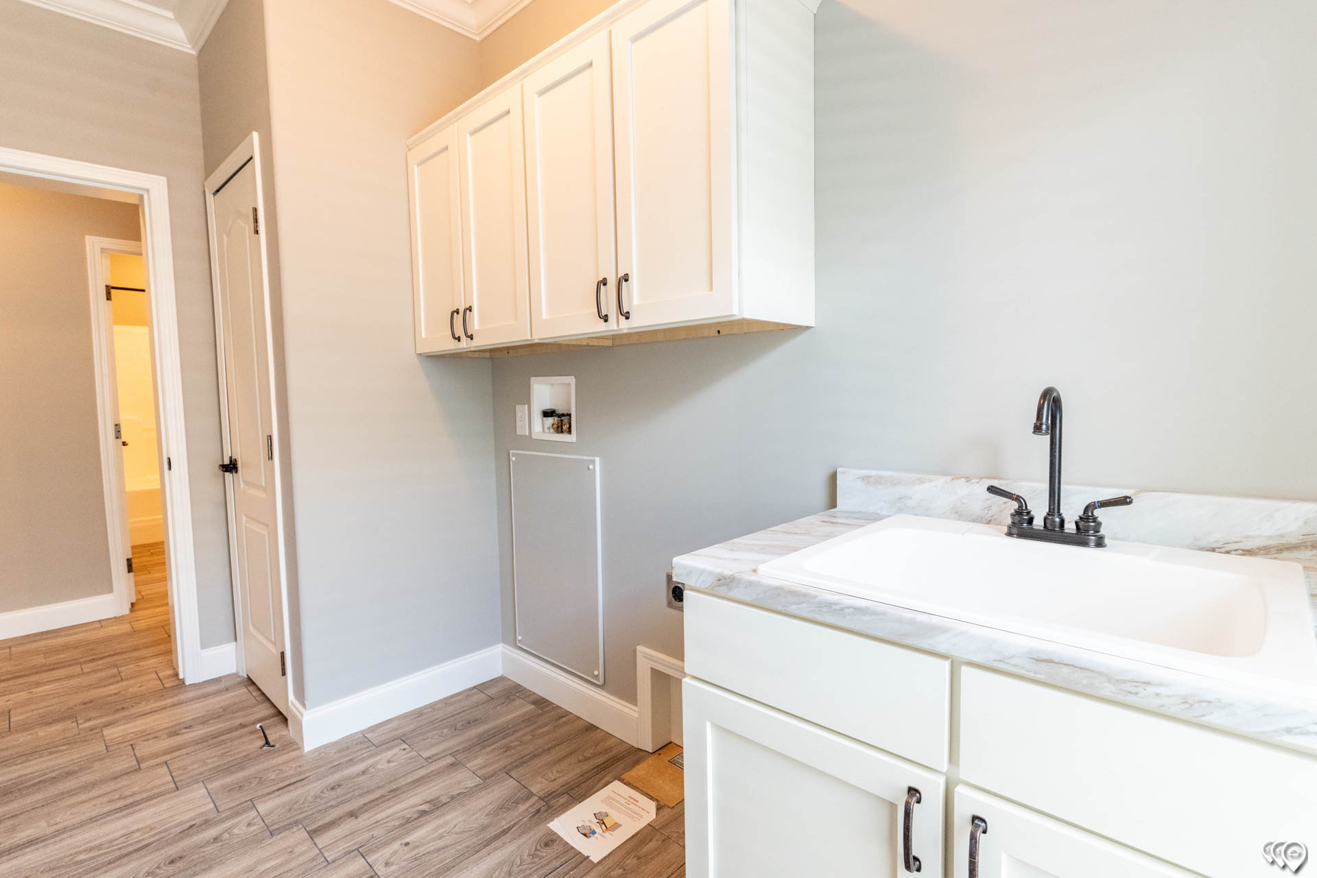 A bright, modern laundry room features light gray walls, wood-textured tile flooring, white cabinets, and a marble countertop with a black faucet.