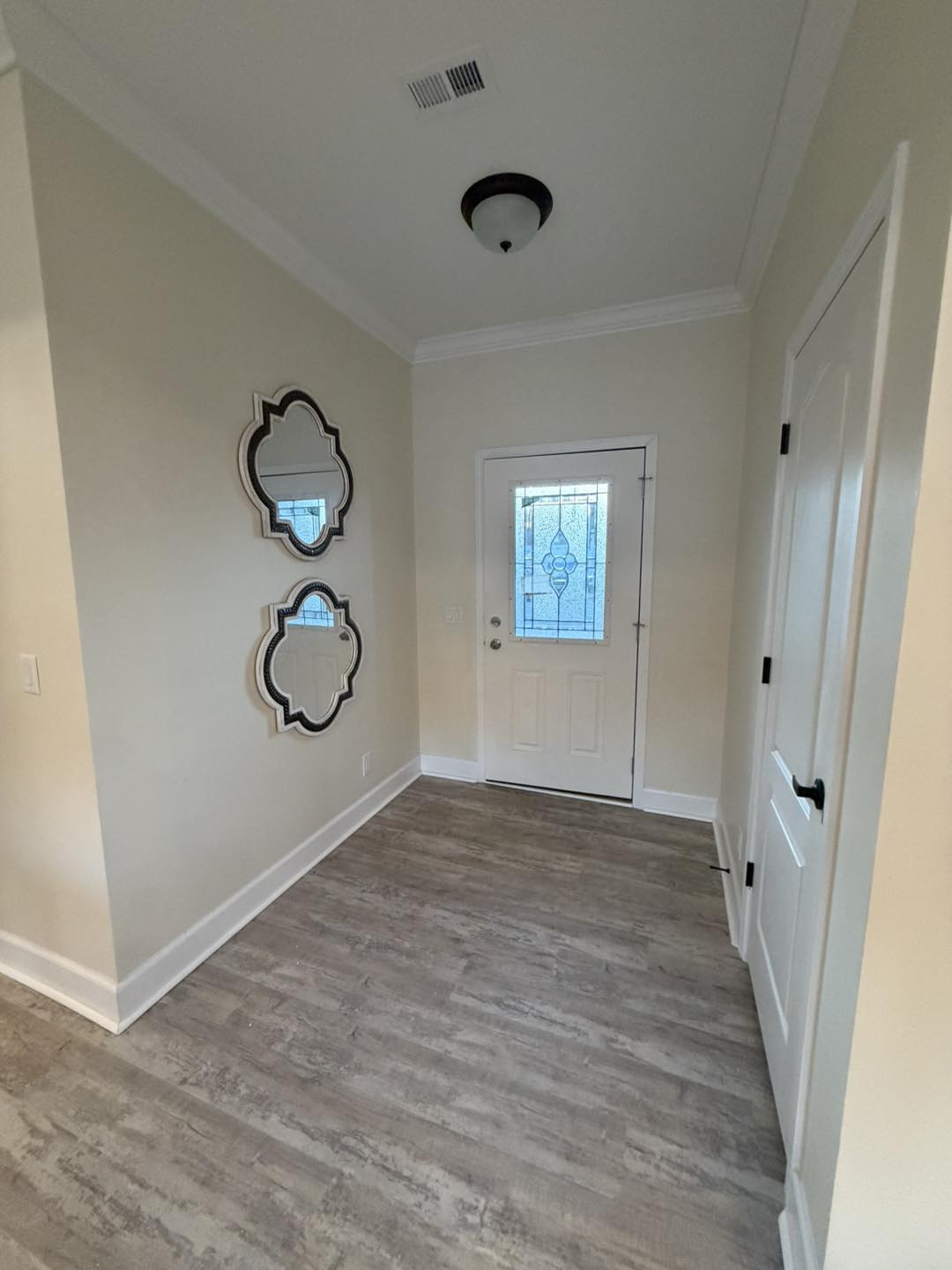 Hallway with light beige walls, wood flooring, and a white door featuring a decorative glass panel. Two ornate mirrors are hung on the left wall.