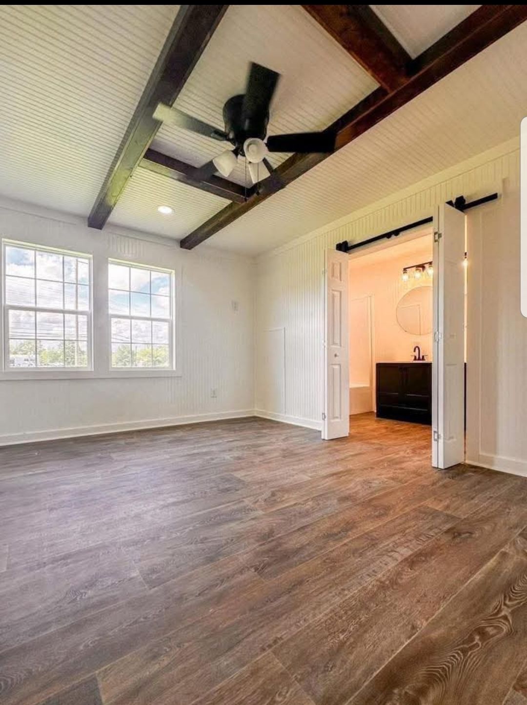 Bright room with wooden floors, white paneled walls, dark wood ceiling beams, and a black ceiling fan. Open door reveals an adjoining bathroom.