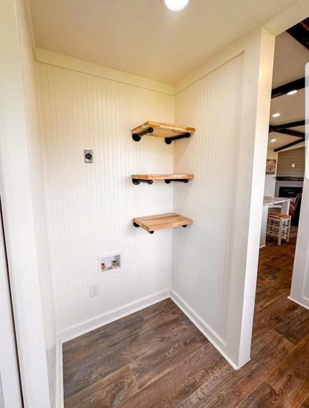 Corner with three wooden shelves on white shiplap walls, industrial pipe supports. Wooden floor adds warmth. Cozy and minimalist interior.