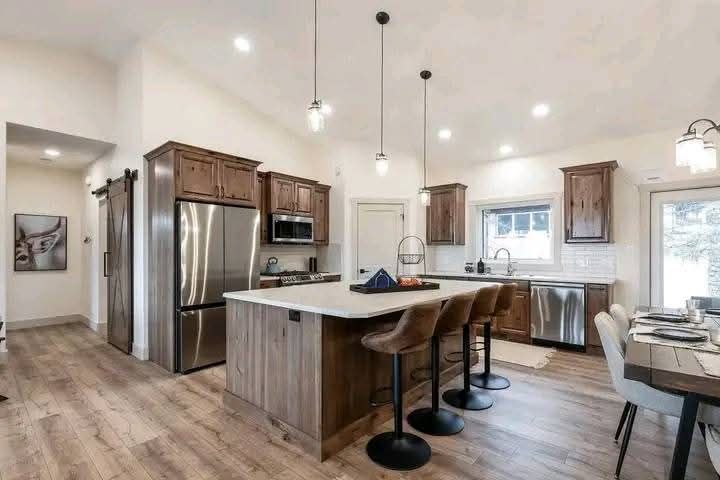 Spacious modern kitchen with wooden cabinets, a central island with four stools, stainless steel appliances, and a dining table. Soft lighting, cozy and inviting.