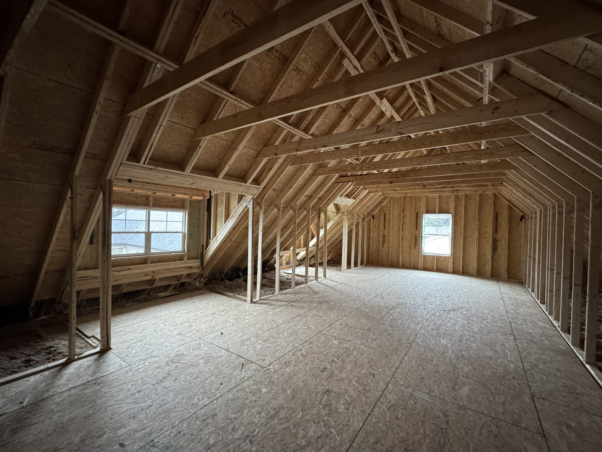 Unfinished attic with wooden framing and plywood flooring. Sloped ceilings and two windows bring in natural light, creating a spacious, airy atmosphere.