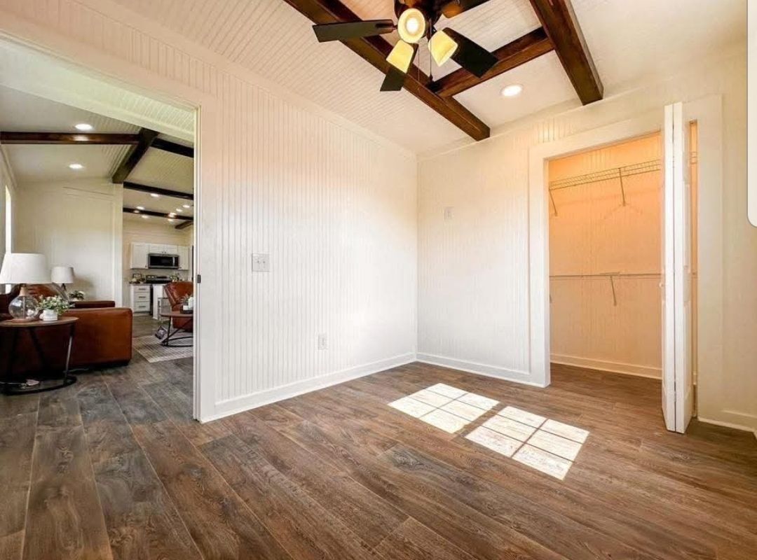 Small room with wooden floor, white walls, and beamed ceiling. Sunlight casts a window pattern on the floor. Open doorway leads to living area. Cozy and inviting.