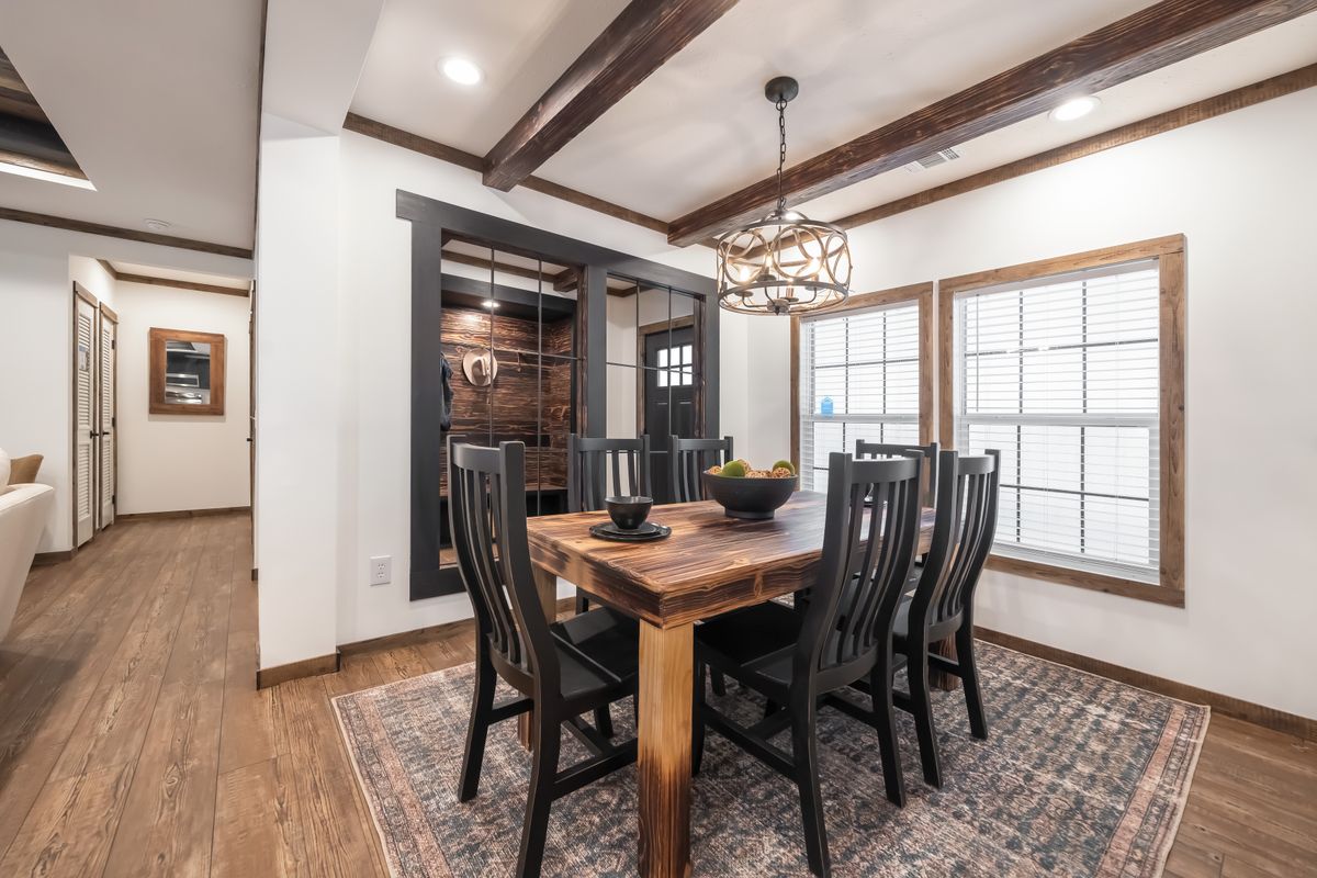 Dining room with rustic wooden table and six black chairs on a patterned rug. Large windows, exposed beams, and a modern chandelier create a cozy ambiance.