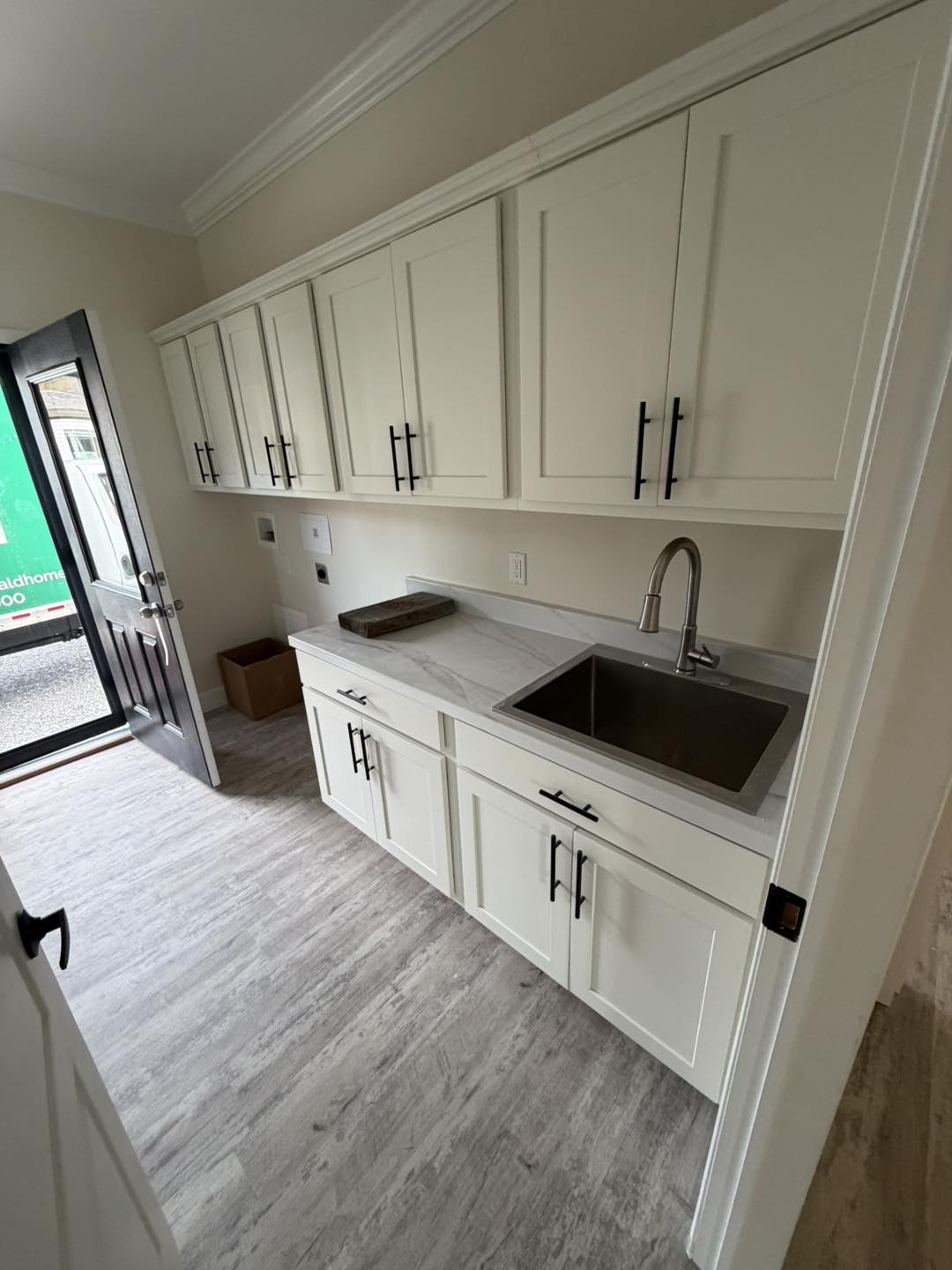 A modern utility room with white cabinets, a silver sink, marble countertop, and light wood flooring. The open door reveals a truck outside.