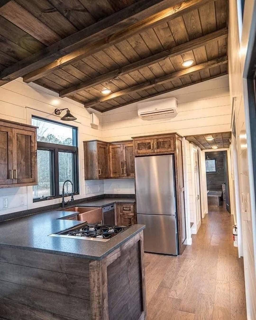 Cozy kitchen with rustic wood cabinets and ceiling. Features a stainless steel fridge, black countertops, and large window. Warm and inviting ambiance.