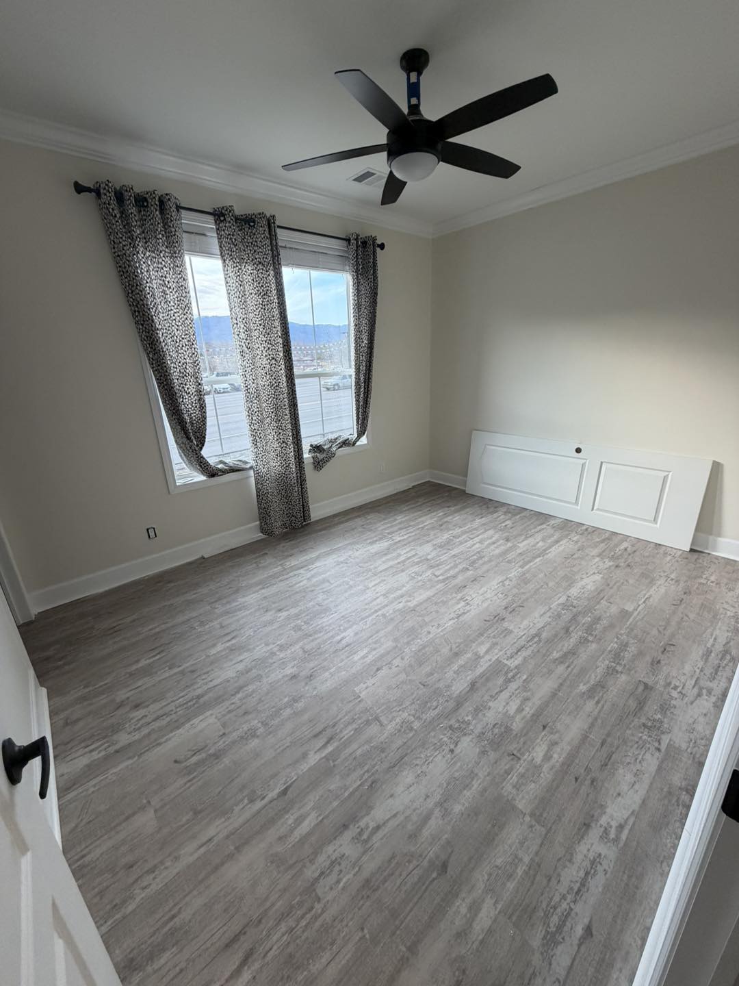 Empty room with light wood flooring, beige walls, and a modern black ceiling fan. A window with leopard print curtains lets in natural light.