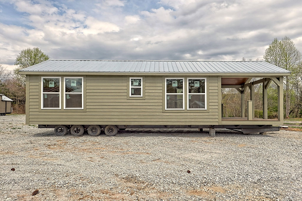 A beige, mobile tiny house on wheels with a metal roof is situated on gravel, featuring large windows and a small porch, under a cloudy sky.