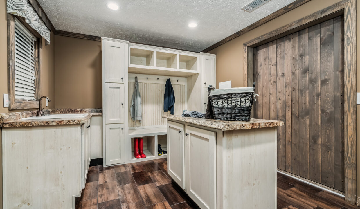 Rustic mudroom with white cabinetry, a wooden bench with hooks, and storage cubbies. A basket sits on the granite-topped island. Warm lighting.