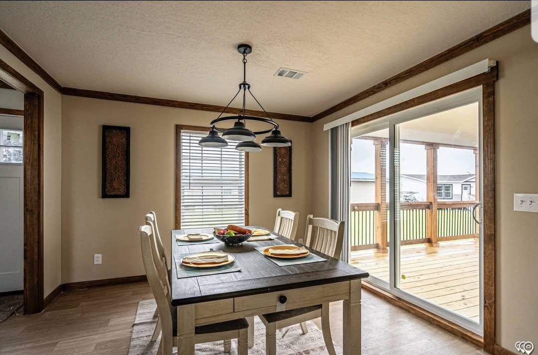 A cozy dining room with a rustic table set for six, featuring light wood chairs and a dark wooden tabletop. A modern light fixture hangs above, and a sliding glass door leads to a porch, creating a bright and inviting atmosphere.