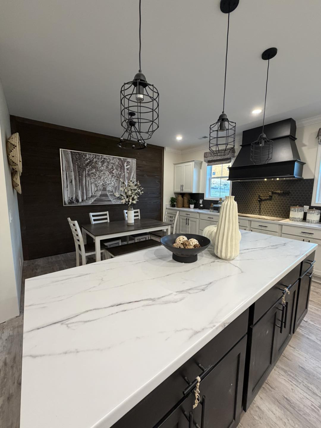 Modern kitchen with a large white marble island, black cabinets, and geometric pendant lights. A dining table and a forest-themed wall art add elegance.