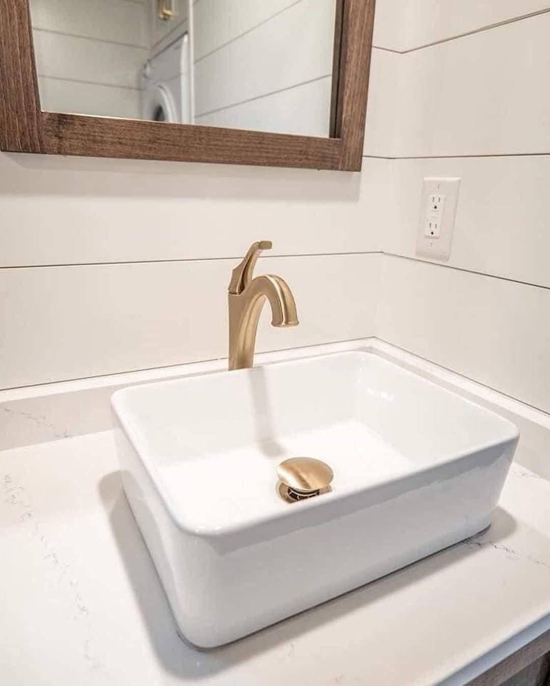 Modern bathroom sink with a rectangular white basin and gold faucet, set against white paneled walls. A wooden-framed mirror is above the sink.