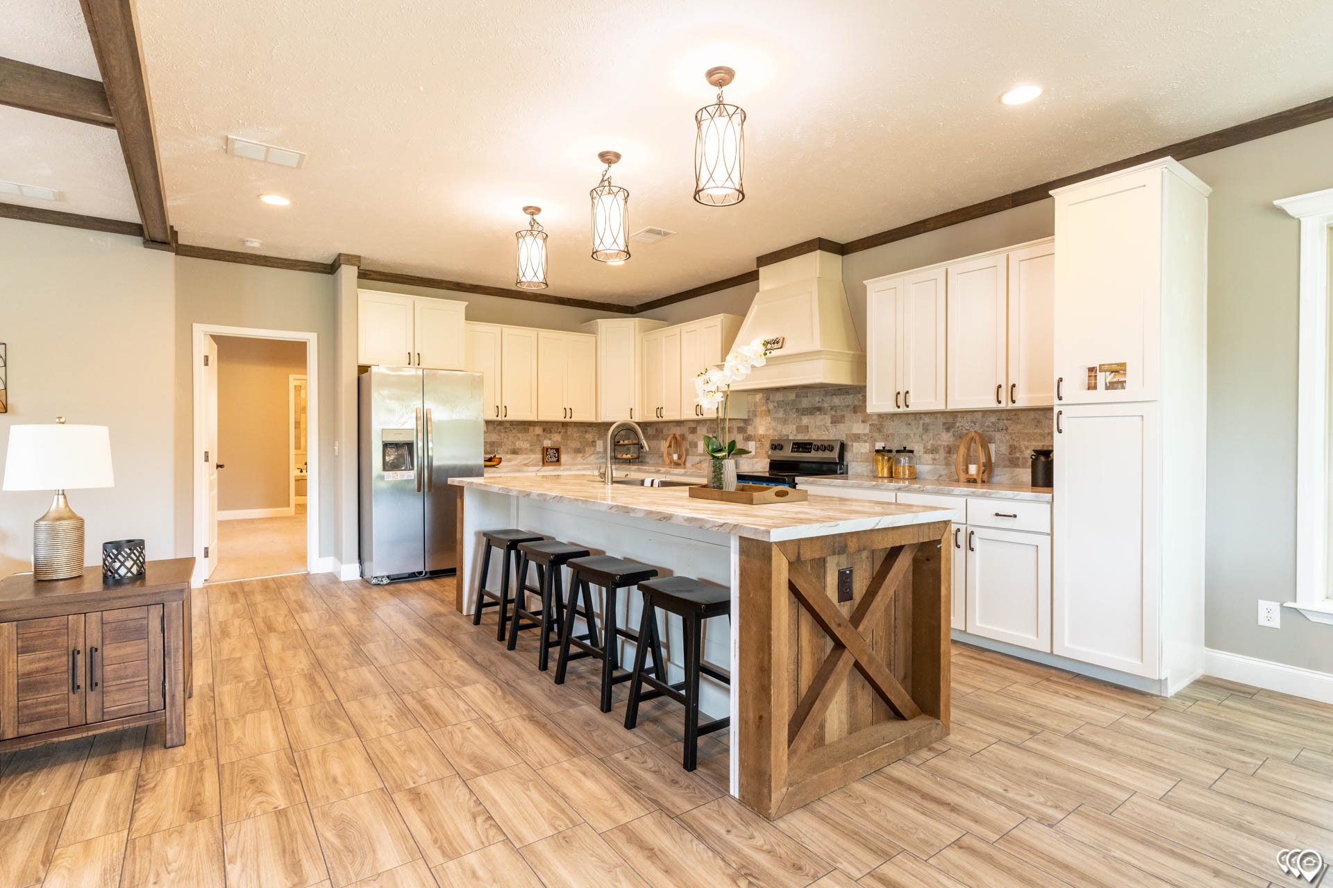 A spacious kitchen with a warm ambiance features white cabinets, a large island with four black stools, modern pendant lights, and stainless steel appliances.
