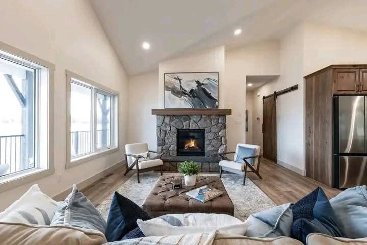 Modern living room with a stone fireplace and abstract art above it. Two armchairs and a cushioned coffee table create warmth. Large windows add brightness.