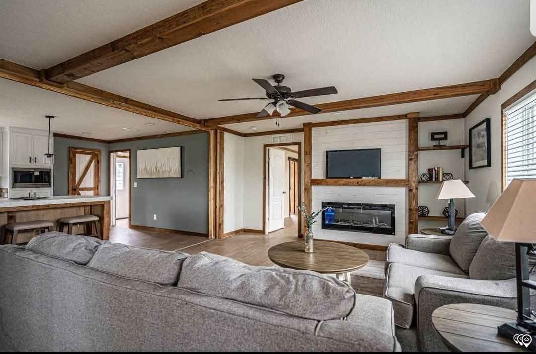 A cozy living room with gray sofas, a round wooden table, and a mounted TV above a fireplace. Exposed wooden beams and neutral tones create a warm, inviting atmosphere.