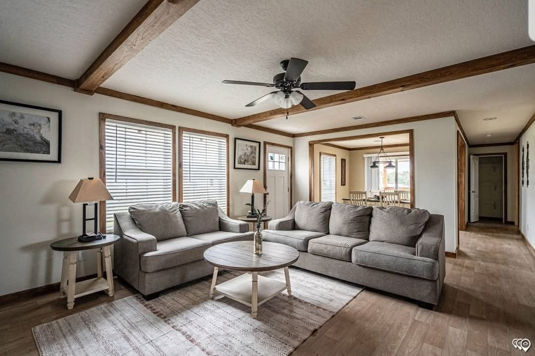 Cozy living room with gray sectional sofa, wooden coffee table, two lamps, and ceiling fan. Light streams through windows, highlighting wooden beams.