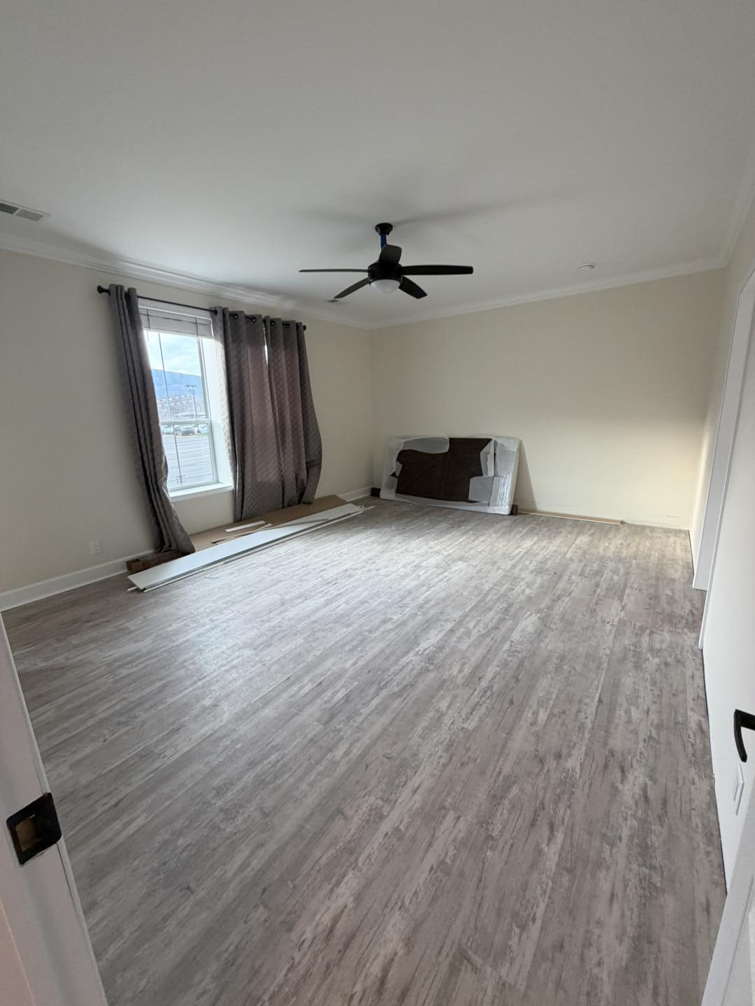 Spacious, empty room with light wood floors, beige walls, ceiling fan, and large window with gray curtains. Unassembled furniture rests against a wall.