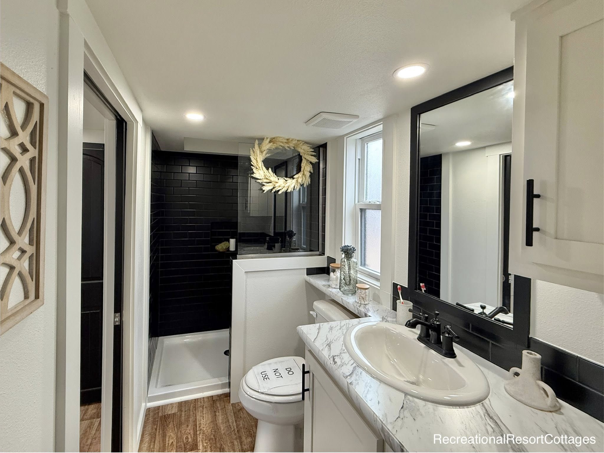 Compact bathroom features a white marble countertop with black fixtures, wood floor, black-tiled shower with a wreath, and a "Do Not Use" sign on the toilet.