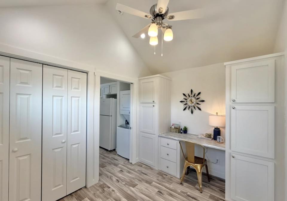Bright white room with a ceiling fan and wood flooring, featuring built-in cabinets. A small desk area with a gold chair is decorated with a lamp and wall art.