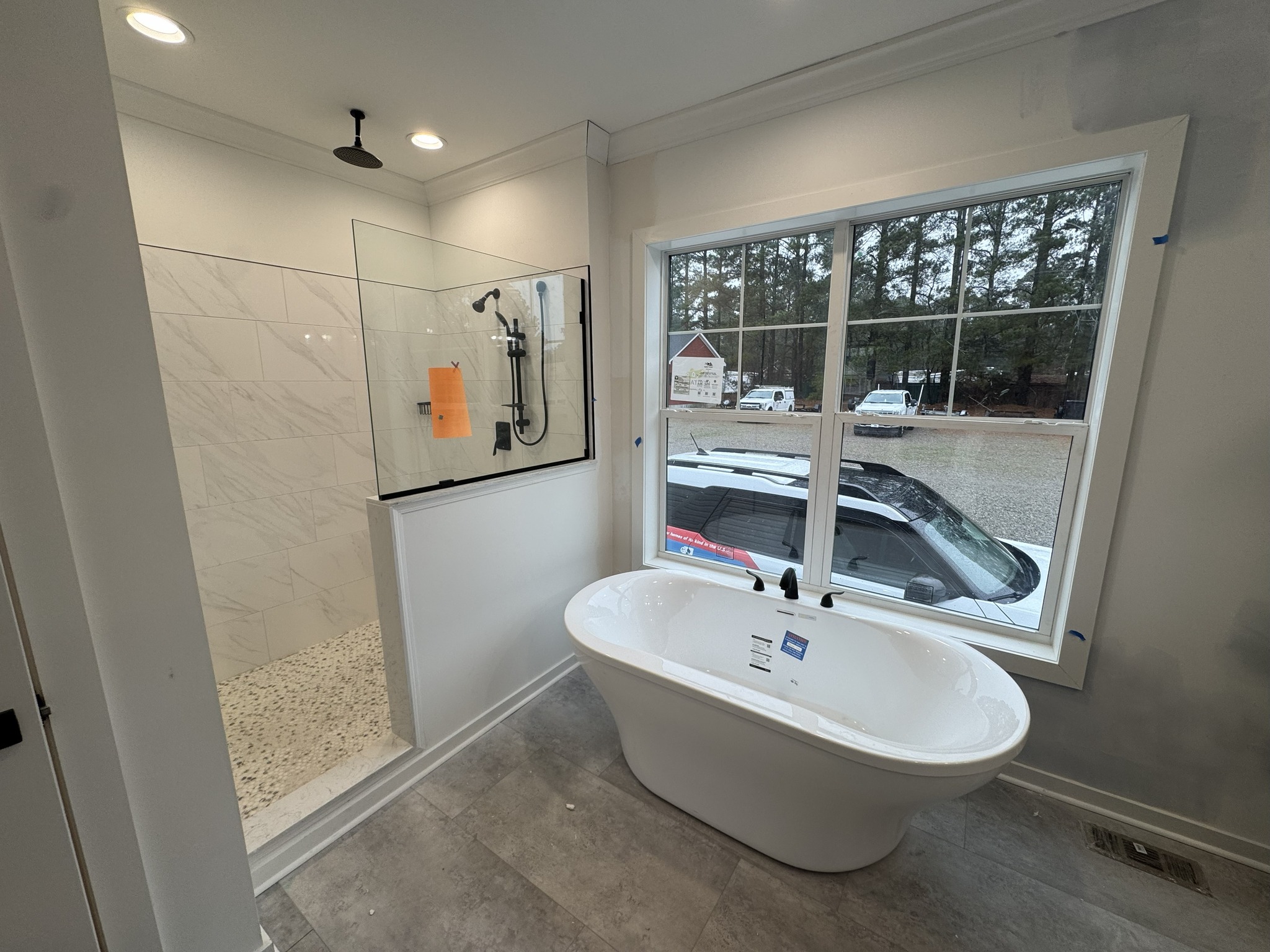 Modern bathroom with a white freestanding tub near a large window showing a driveway and trees. A glass-enclosed shower is on the left with a black showerhead.