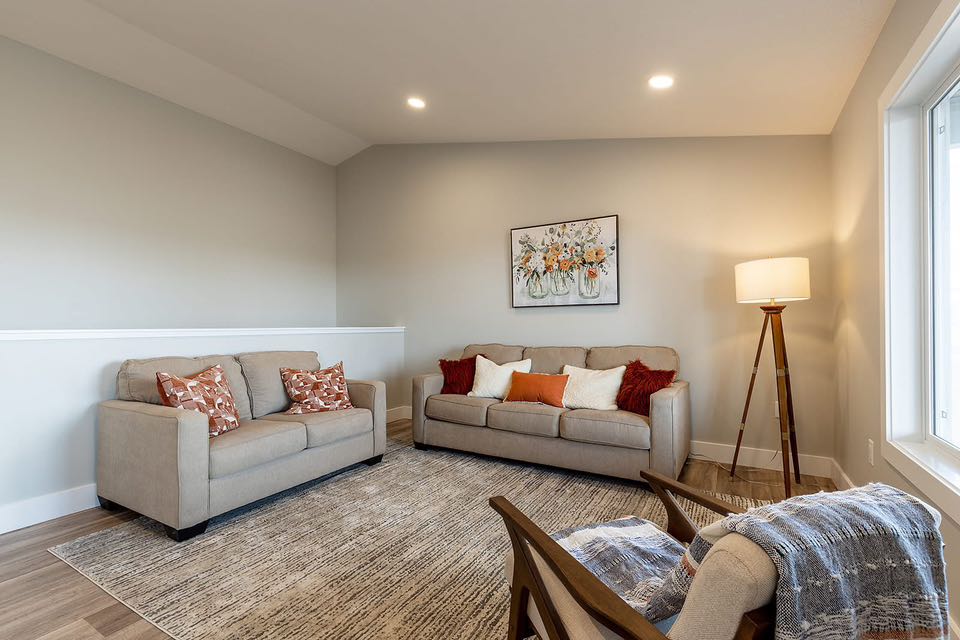 Cozy living room with beige sofas, colorful cushions, and a floor lamp. A floral painting adorns the wall. Warm, inviting atmosphere.