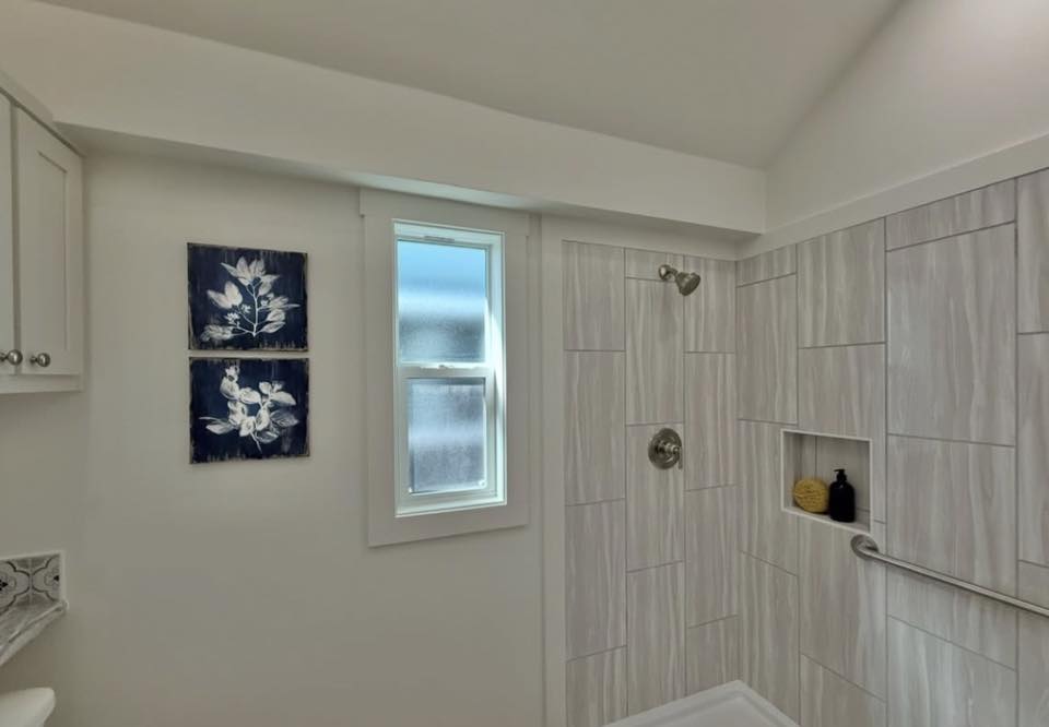 Modern bathroom with light gray tiled shower. A window, floral wall art, and built-in niche with bottles add elegance. Bright, clean atmosphere.