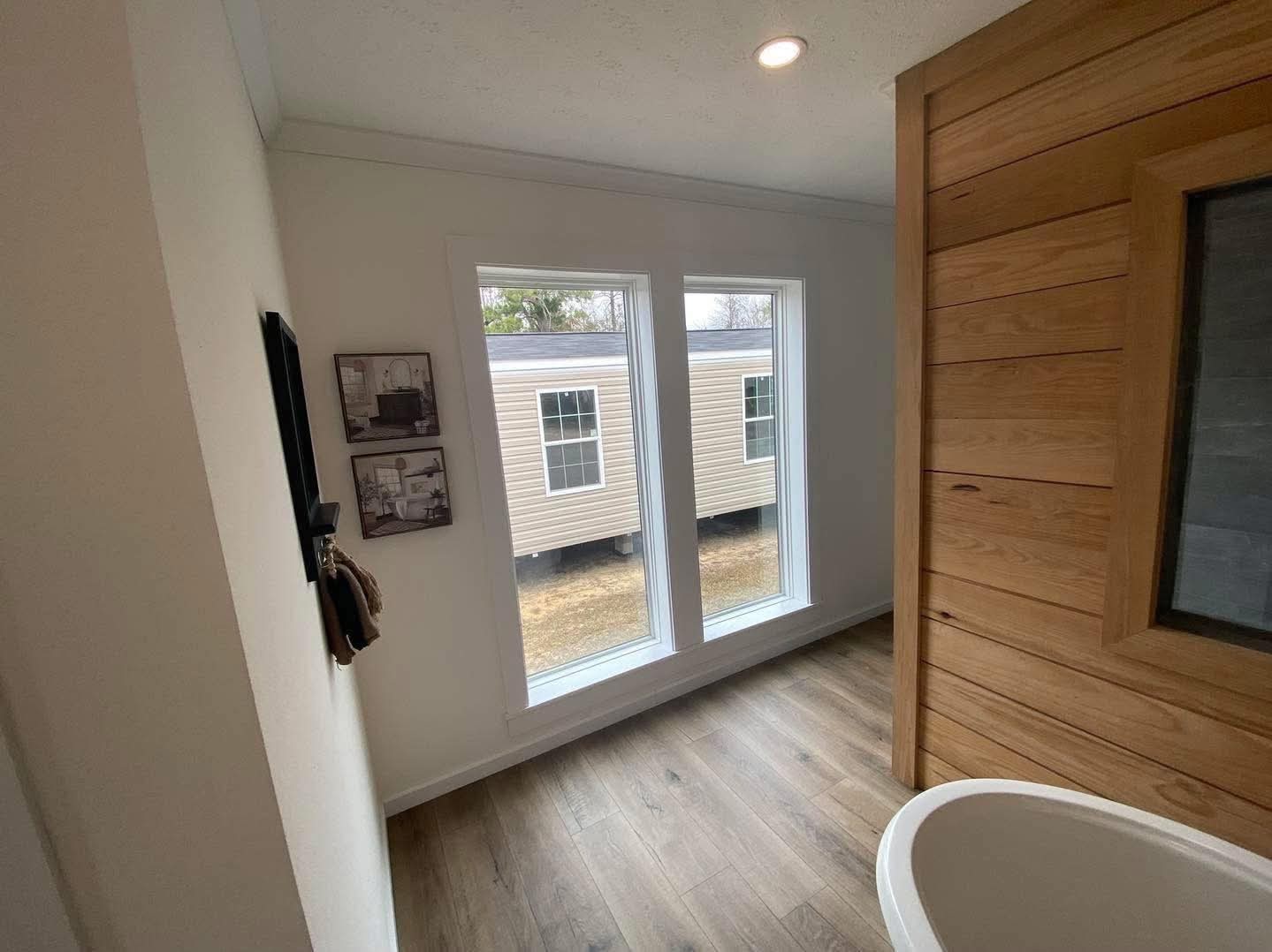 Bright bathroom with large windows showing an exterior view of another building. A wooden accent wall adds warmth, creating a calm, inviting atmosphere.